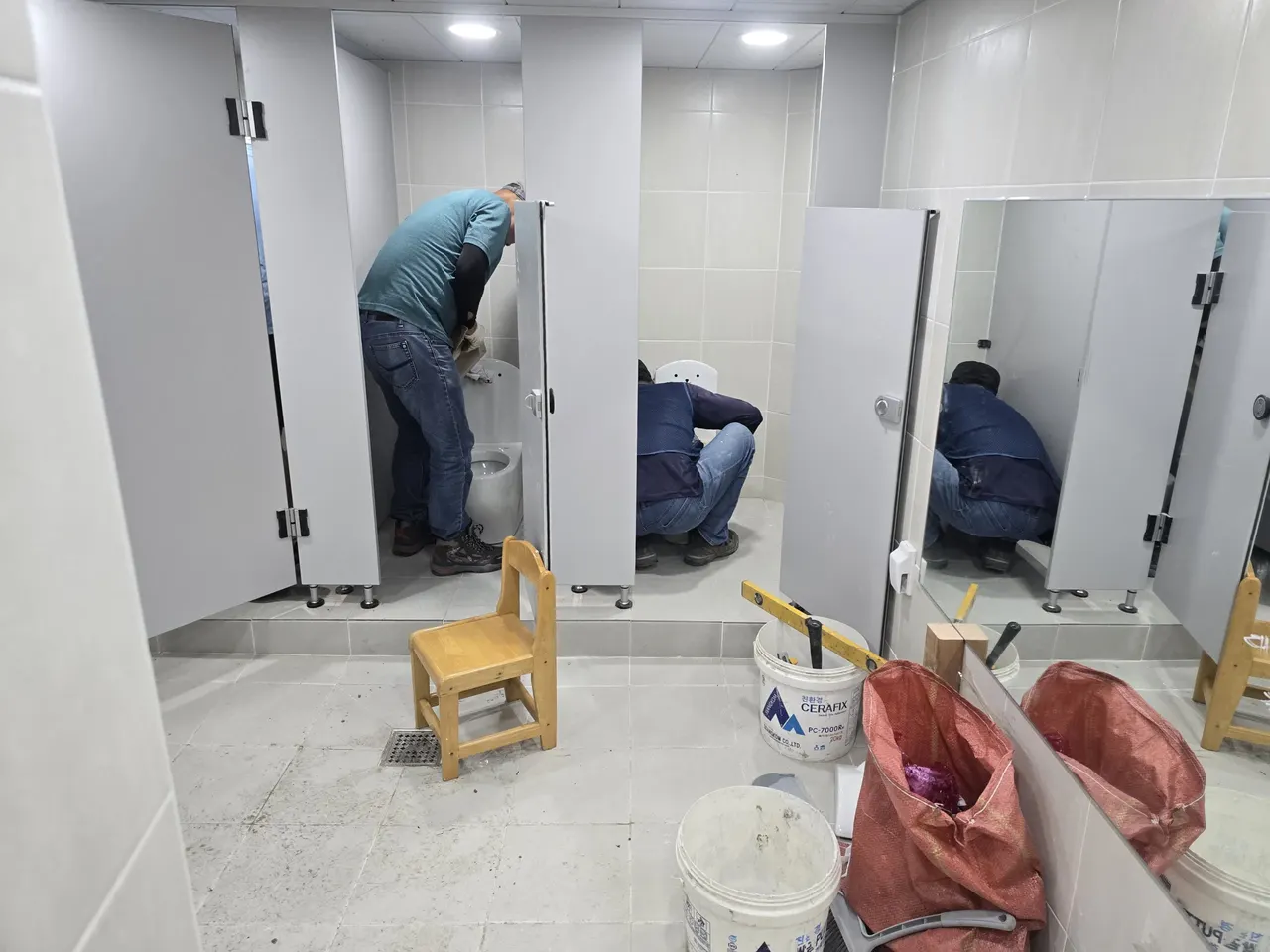 화장실 변기 칸막이 내부에서 작업자들이 몸을 숙이고 작업하는 모습, 옆에 시멘트 통과 타일 자재