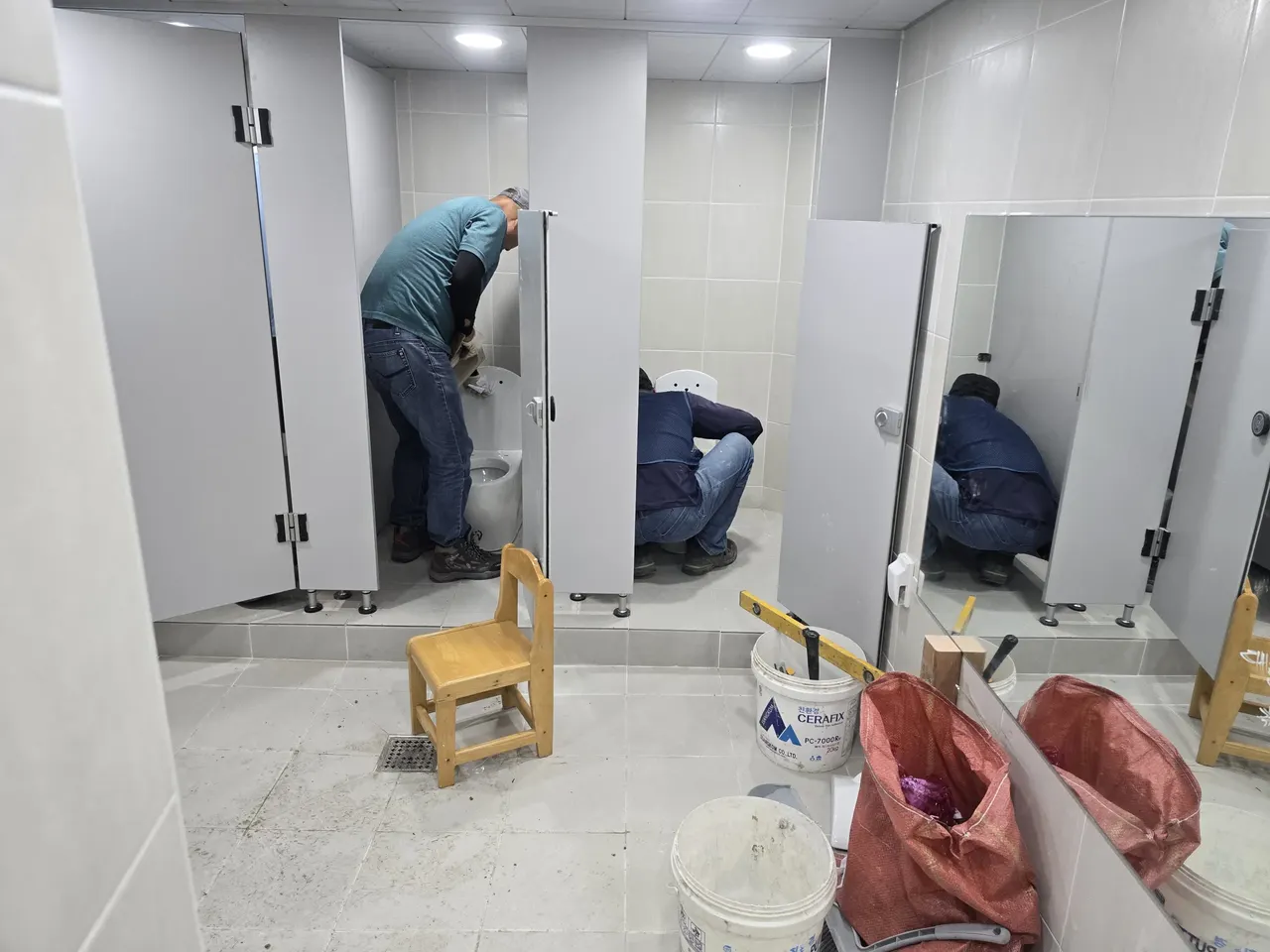 화장실 변기 칸 여러 곳에서 작업자들이 등을 숙여 작업하는 현장, 시멘트 통과 타일 마감재 포대